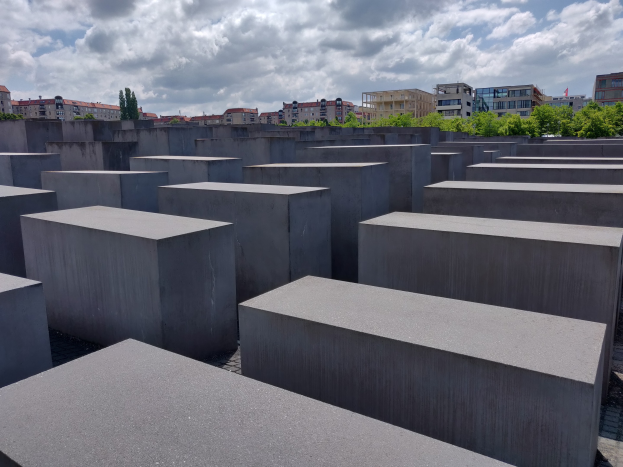 Betonschachteln in einem Gittermuster am Holocaust-Mahnmal für die ermordeten Juden Europas in Berlin, mit Bäumen, Gebäuden und Wolken im Hintergrund.