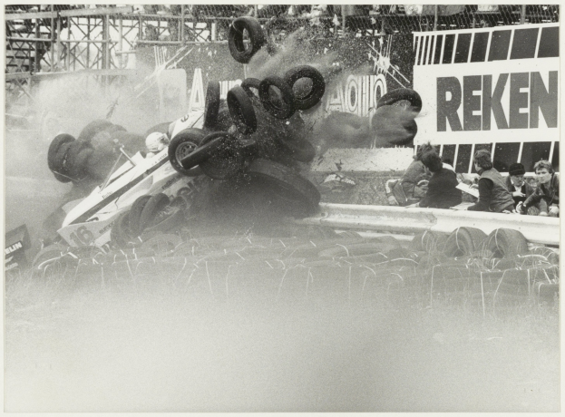 Schwarzes und weißes Foto eines zertrümmerten Rennwagens, umgeben von Reifen, mit Menschen zu seiner Rechten und einer Tafel mit Text im Hintergrund.