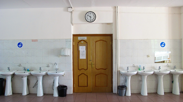 Ein Badezimmer mit einer Reihe von Waschbecken, Bodenschmutzbehältern, einer Tür, einer Wanduhr und einem Spiegel auf der rechten Seite.