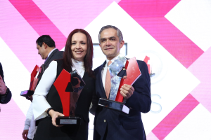 Zwei Personen auf einer Bühne mit Trophäen stehend und strahlend, mit einem Bildschirm im Hintergrund.