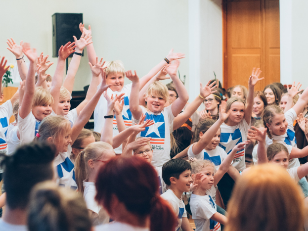 Gruppe von Kindern, die Hände vor einer Menge heben, mit einem Tisch, einer Vase, einem Lautsprecher, einer Wand und einer Tür im Hintergrund.