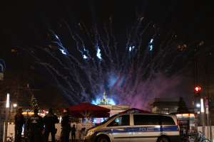 Ein Polizeiwagen steht vor einer Menschenmenge, umgeben von Fahrrädern, Zelten, Laternenmasten, Schildern, Verkehrszeichen, Bäumen, Gebäuden und einer Statue im Hintergrund, unter einem von Feuerwerk erhellten Himmel.