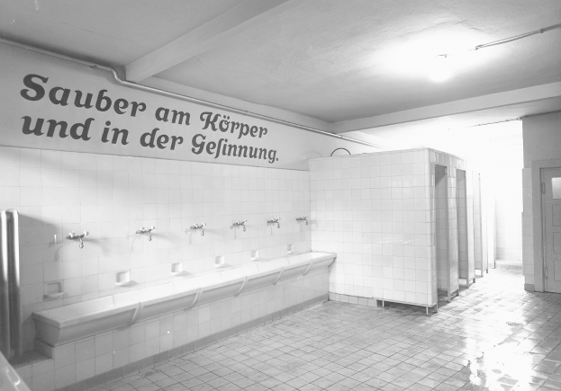 Schwarz-weißes Foto einer öffentlichen Toilette mit Fliesenboden, einer Wand mit Text, mehreren Wasserhähnen, einer Tür und einer Deckenlampe.
