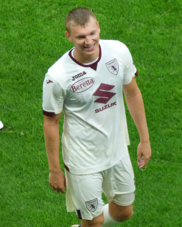 Ein Mann in einem weißen Hemd und Shorts lächelt beim Laufen auf einem Fußballfeld, trägt Schuhe.