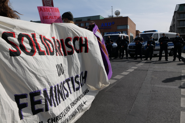 Eine Gruppe von Menschen marschiert auf einer Straße, hält eine Fahne mit der Aufschrift "Solidarität und Feminismus" und hat parkende Fahrzeuge und Gebäude im Hintergrund unter einem klaren blauen Himmel.
