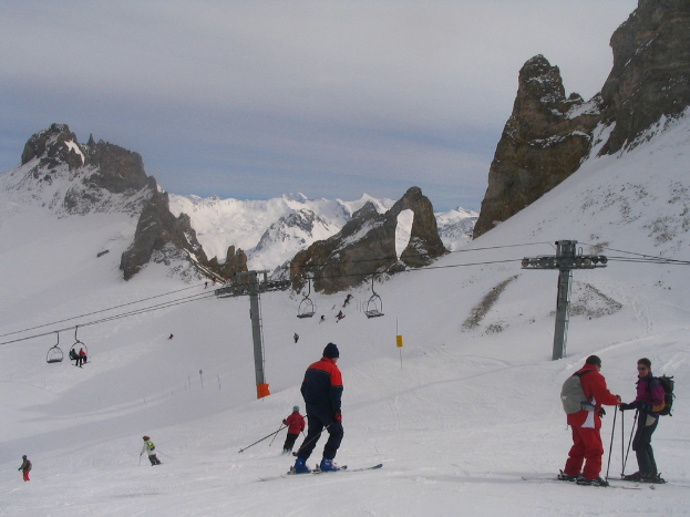 Menschen in Pullovern fahren mit Skiern auf dem Eis und einer Seilbahn, Bergen und einem bewölkten Himmel im Hintergrund.