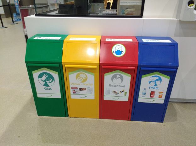 Vier bunte Mülltonnen in einer Reihe auf dem Boden mit einer Theke, Monitor und anderen Gegenständen im Hintergrund, wahrscheinlich zeigt Recycling-Behälter in Singapur.
