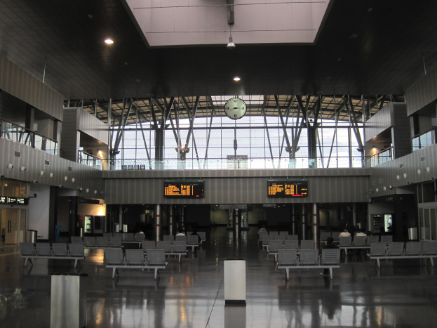 Ein leerer Flughafen mit zahlreichen leeren Stühlen und Tischen, verstreuten Mülltonnen, Informationsanzeigen, Deckenleuchten, Stützstangen, einer Uhr und Glaswänden im Hintergrund.