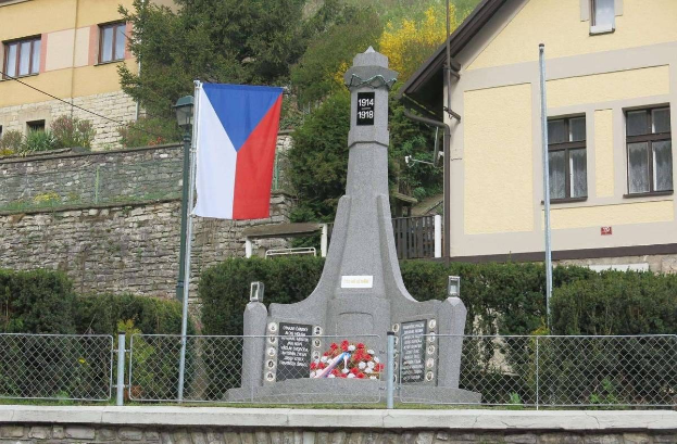 Ein Denkmal mit einer Flagge darauf, umgeben von einem Zaun, Pflanzen, Bäumen und einer Wand, vor einem Gebäude, gewidmet denen, die während des Zweiten Weltkriegs ihr Leben verloren haben, mit eingravierter Inschrift.