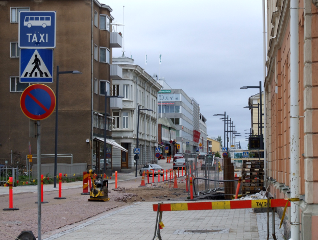 Eine Stadtstraße mit Gebäuden, Straßenlaternen, Verkehrsschildern, Verkehrskegeln, Kraftfahrzeugen, Absperrpoller, Bäumen und einer Baustelle mit Verkehrsschildern unter einem bewölkten Himmel.