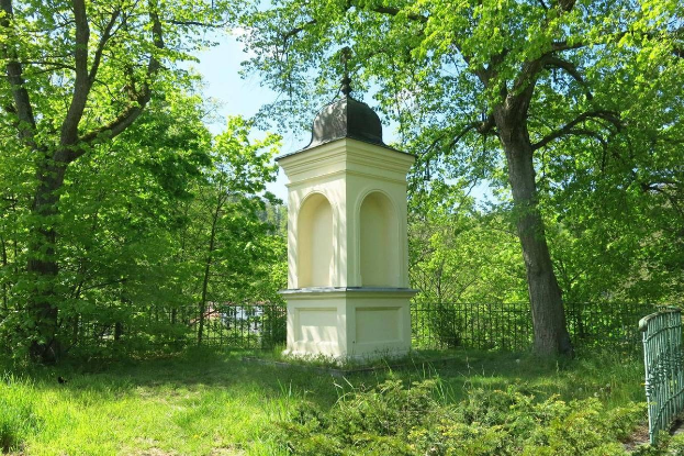 Kleines weißes Holocaust-Gedenkmal auf einem Rasenfriedhof umgeben von einem Zaun und Bäumen unter einem klaren blauen Himmel in Vilnius, Litauen.