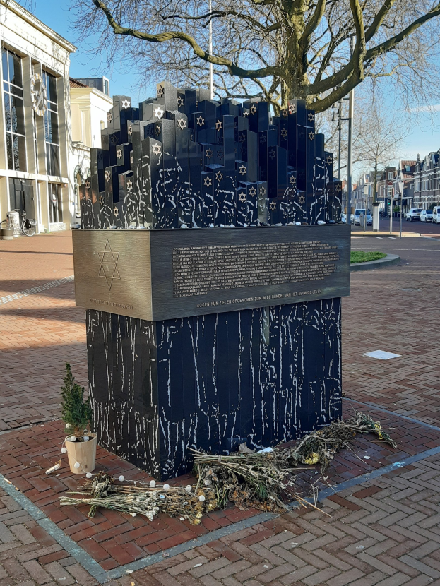 Denkmal in einem Stadtplatz gewidmet an Holocaust-Opfern, mit einem Baum davor, einem Topfgewächs, verstreuten trockenen Blättern, umliegenden Gebäuden, Straßenverkehr, Strommasten und Bäumen unter einem Himmel.