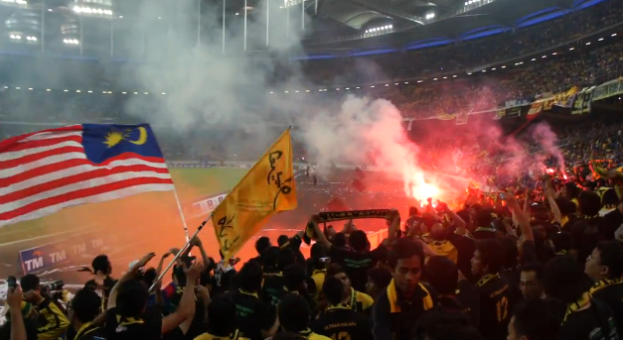 Eine große Menge in einem Stadion mit Fackeln, Fahnen und einer Kamera, mit Rauch und Schildern mit der Aufschrift "Malaysia vs Malaysia Live Streaming" im Hintergrund.