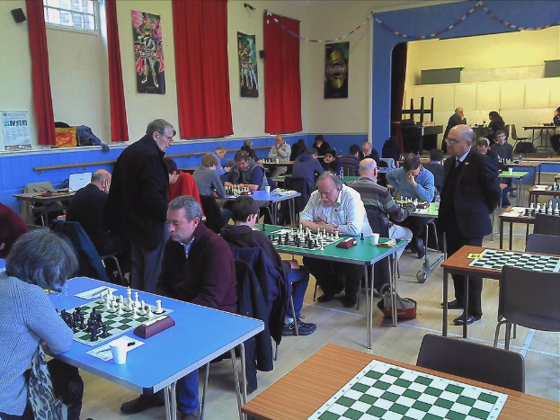 Zwei Personen spielen Schach an einem Tisch in einem großen Saal mit Vorhängen, Plakaten, einem Fenster und einem Kalender an der Wand.