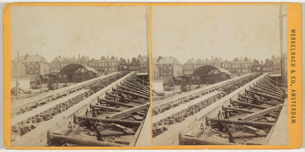 Ein altes Schwarz-Weiß-Foto einer Bahnschiene mit Gebäuden im Hintergrund, Text auf der rechten Seite, der 'Merkelbach & Co, Co. Amsterdam' lautet, und verstreute hölzerne Gegenstände im Vordergrund.