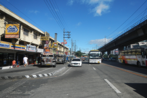 Eine belebte Stadtstraße mit Autos, Bussen, Fußgängern, Gebäuden, Strommasten, einer Brücke, Bäumen und einem bewölkten Himmel.