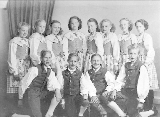 Schwarzes und weißes Foto einer Gruppe von Kindern, die vor einer Wand mit einem Vorhang im Hintergrund posieren, mit der Beschriftung "1930er Kinder-Schule" unten.