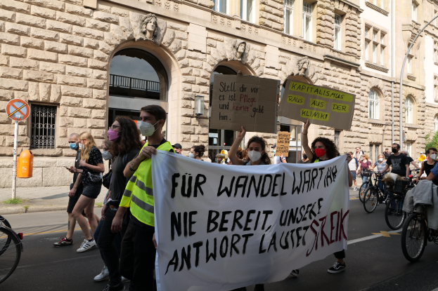 Eine Gruppe von Menschen marschiert auf einer Straße in Berlin, hält Schilder und Banner hoch und fährt mit Fahrrädern, während sie gegen die Anti-Kriegsbewegung protestieren, mit einem Gebäude im Hintergrund, das Fenster, Bögen, Säulen, Skulpturen, Bäume und einen Laternenmast aufweist.