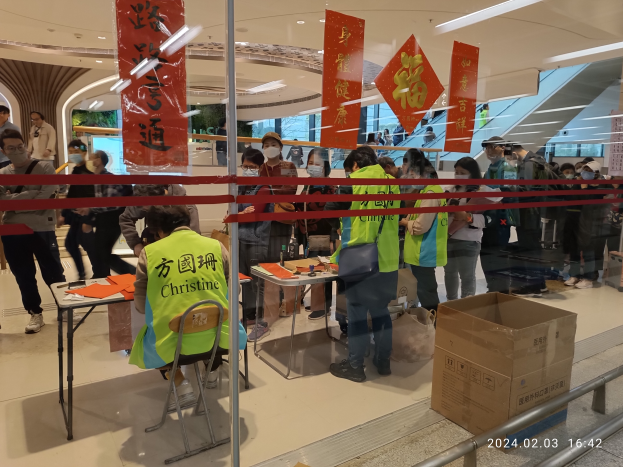 Eine Gruppe von Menschen mit Mund-Nasen-Schutz steht an einem Tresen in einem Einkaufszentrum an. Es sind Stühle und Tische mit verschiedenen Gegenständen darauf zu sehen, und eine Person sitzt auf einem der Stühle. Vor dem Tresen befindet sich ein Geländer, und von der Decke hängen Schilder mit Text. Rechts im Bild ist ein Karton und ein Wasserzeichen zu sehen.