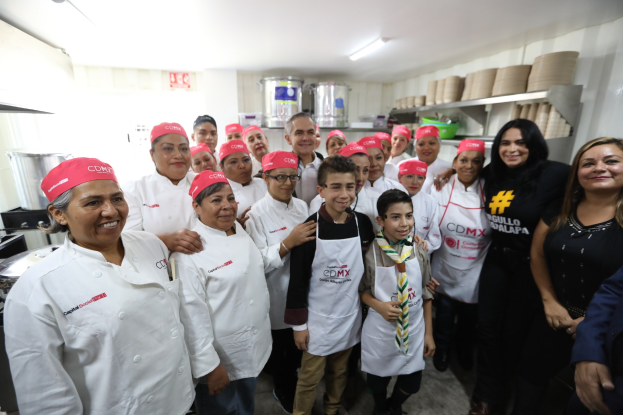 Eine Gruppe von Menschen in einer Küche, die l├Ąchelt und für ein Foto posiert, einige tragen Küchenhüte, mit Utensilien links und Küchenregalen im Hintergrund.