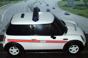 Ein weißer Polizeiwagen parkt vor einer befahrenen Straße mit fahrenden Fahrzeugen, umgeben von Gras und Bäumen, in einer ländlichen Umgebung.