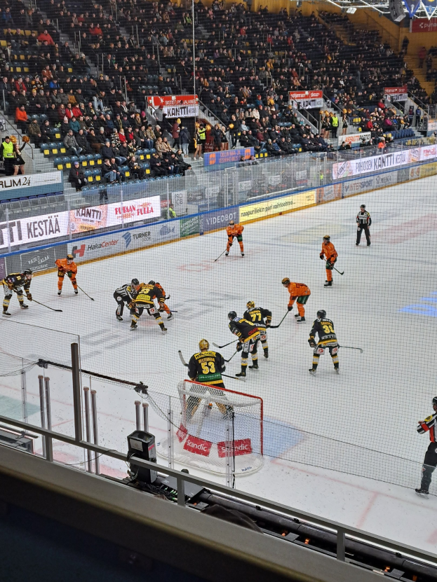 Gruppe von Menschen, die Hockey auf einem Eisstadion spielen, umgeben von einem Zaun, mit einem Netz in der Mitte, Zuschauern im Hintergrund, Bannern mit Text und Stadionbeleuchtung von oben.