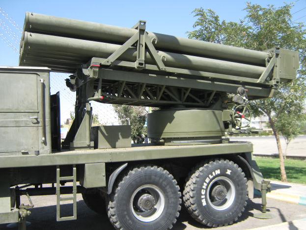 Ein militärisches Lastwagenfahrzeug mit einer Rakete auf der Ladefläche fährt eine Straße entlang, umgeben von Gras, Bäumen, Gebäuden und einem Maschendrahtzaun.