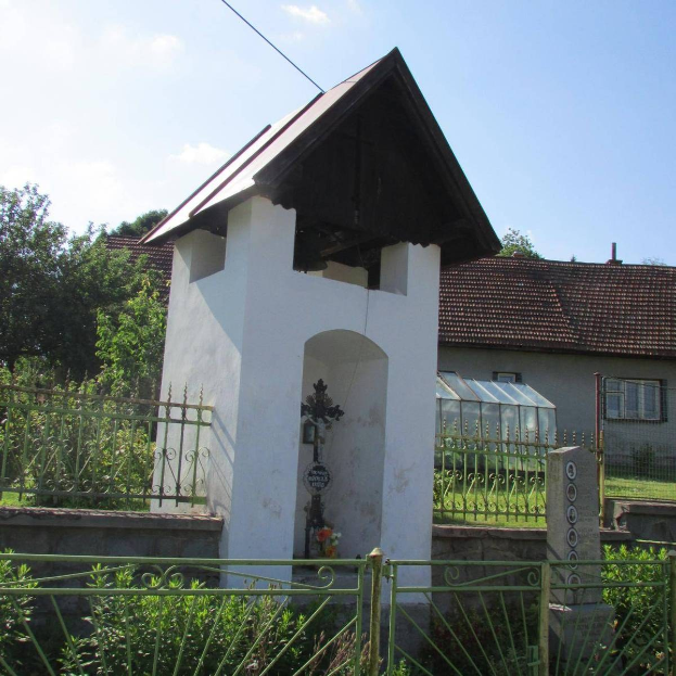 Außenansicht der Heilig-Geist-Kirche mit einem kleinen weißen Gebäude mit einem Kreuz, einem Metallzaun, umgeben von Grün, nahegelegenen Häusern und einem bewölkten Himmel.