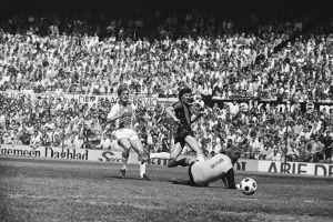 Schwarzes und weißes Foto von Fußballspielern im Einsatz auf einem Feld mit Zuschauern und einem Bundesliga 1978-79-Spielbanner im Hintergrund.