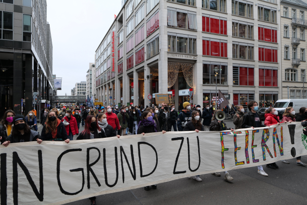 Eine Gruppe maskierter Personen hält ein Banner mit der Aufschrift "In Grund zu Feiern" vor Gebäuden, Laternenmasten und Fahrzeugen, was auf eine Demonstration in Berlin, Deutschland, hindeutet.