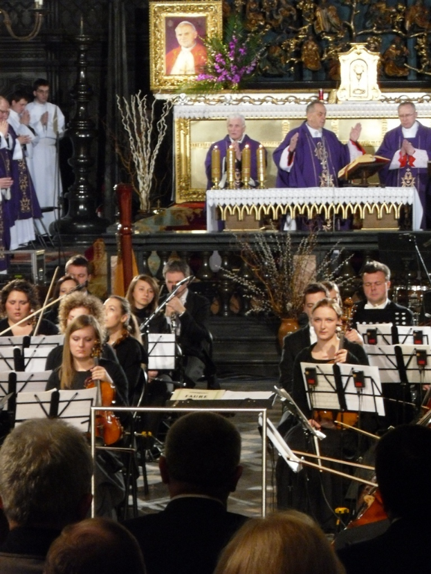 Eine Gruppe von Menschen, die Geigen spielen, mit Noten vor sich, mit drei Kirchenvätern im Hintergrund, einer Bibel davor und einem Photo-Rahmen mit Verzierungen drumherum.