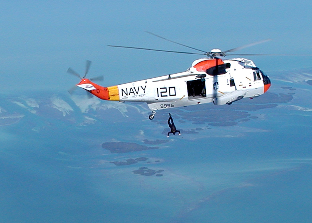 Ein Rettungshubschrauber fliegt über einen Gewässer mit einer Person in der Luft, vor dem Hintergrund des Himmels.