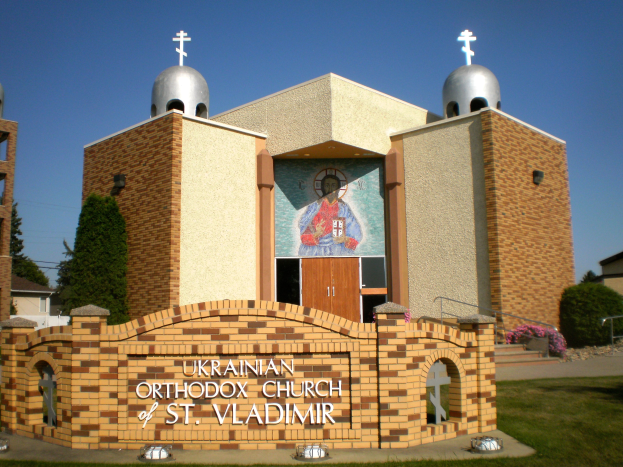 Außenansicht der Ukrainischen Orthodoxen Kirche von St. Wladimir, mit einer textbedeckten Wand, Rasen, umgeben von Gebäuden, Bäumen, blühenden Pflanzen, Treppen mit Geländern und einem Wandgemälde, mit dem Himmel im Hintergrund.