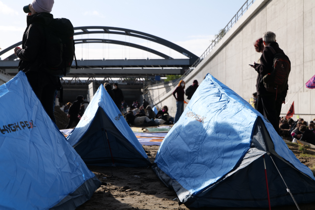 Eine Gruppe von Menschen sitzt auf einem sandigen Strand in der Nähe von Zelten, mit einer Wand auf der rechten Seite und einer Brücke im Hintergrund, die Gegenstände halten und Taschen tragen, was auf eine Klimaprotest unter einem bewölkten Himmel hinweist.