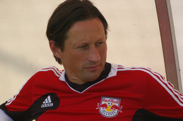 Mann in rot-schwarzem T-Shirt sitzt auf einer Bank vor einer Wand und sieht aus wie ein professioneller Fußballspieler mit entschlossener Miene.