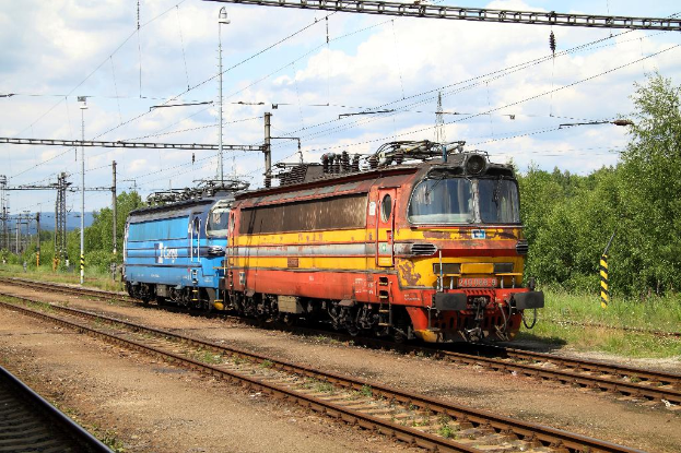 Eine Zugmaschine auf Schienen mit elektrischen Masten, Kabeln, Bäumen, Pflanzen und Wolken am Himmel.