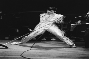 Elvis Presley in weißer Bühnenkleidung bei einem Auftritt im Apollo Theater in Las Vegas, mit Mikrofon in der Hand und einem Musiker rechts daneben mit einem Musikinstrument, mit Bühnenbeleuchtung und -instrumenten im Hintergrund.