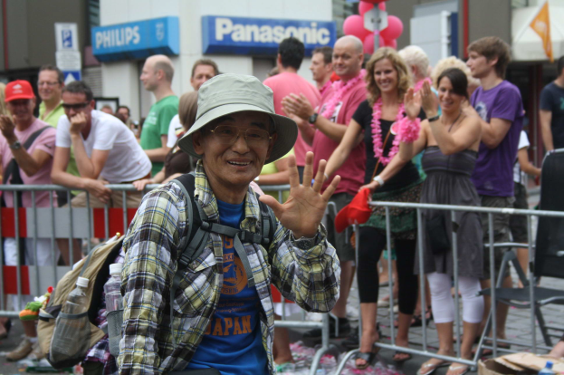 Ein Mann mit Hut, Sonnenbrille und Brille steht vor einer Menge bei einer Gay Pride Parade, mit einigen klatschenden Menschen und einer Absperrung dahinter sowie Gebäuden mit Namensschildern und Ballons im Hintergrund.