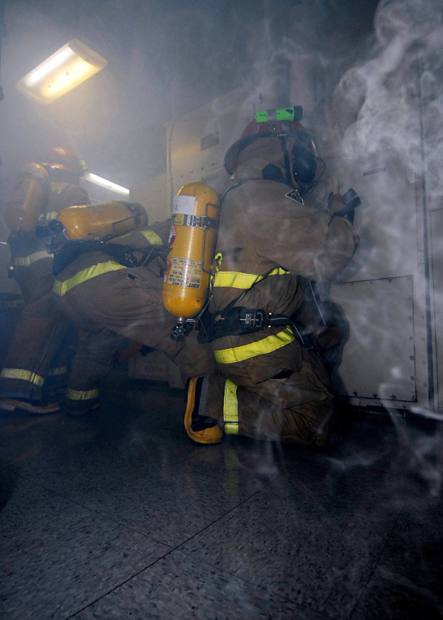 Feuerwehrleute in einem raucherfüllten Raum mit beleuchteter Ausrüstung und einer sichtbaren Wand im Hintergrund.