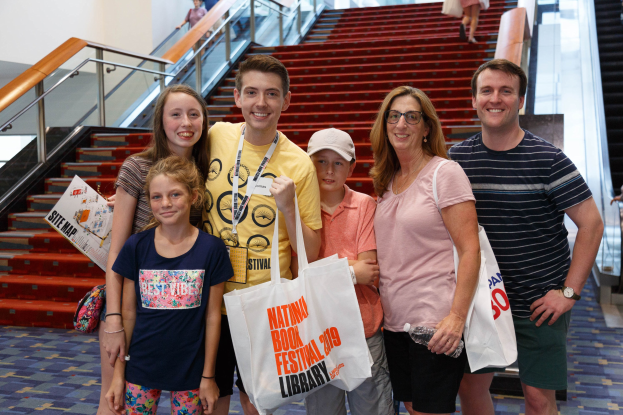 Eine Gruppe von Menschen, die vor einer Rolltreppe stehen und Taschen halten, vor einer Treppe, einem Geländer und einer Wand im Hintergrund.