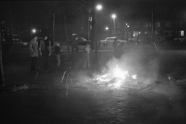 Gruppe von Menschen um ein Feuer auf der Straße bei Nacht versammelt, mit Fahrzeugen und Gebäuden im Hintergrund.