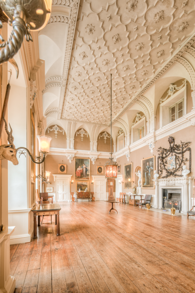 Ein prächtiger Schloss-Saal mit Holzboden, Kronleuchtern, Tischen, Stühlen, Kamin, Statuen, Fotorahmen, Fenstern und diversen anderen Gegenständen.