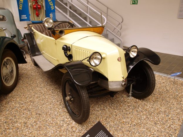 Ein Oldtimer wird in einem Museum ausgestellt, umgeben von Infotafeln, Treppen mit Geländern, Bannern mit Text und Bildern und einer Wand mit einem Fotorahmen.