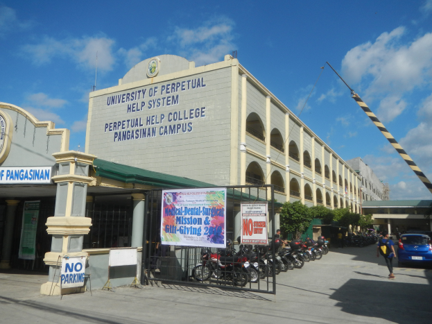 Die Universität Perpetual Help System in Pangasinan mit ihren Gebäuden, geparkten Motorrädern, einem Metallzaun, einer Fahne, Schildern, Menschen, Fahrzeugen, einem Kran, Bäumen und einem bewölkten Himmel.
