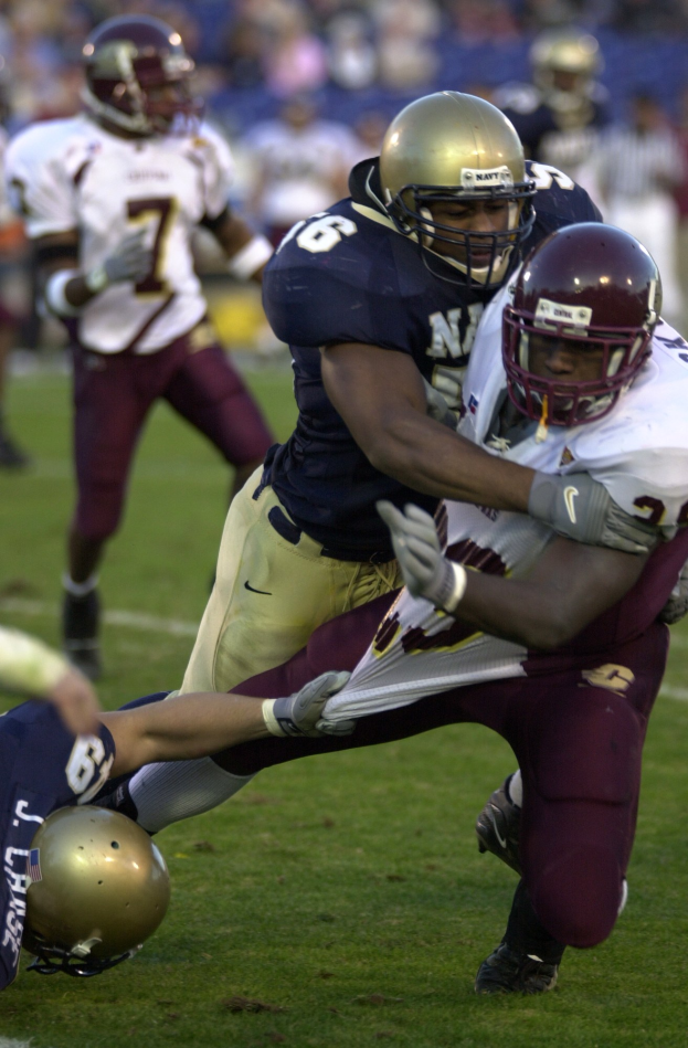 Zwei Fußballspieler in Helmen und Uniformen ringen auf dem Feld, während andere im Hintergrund zusehen.