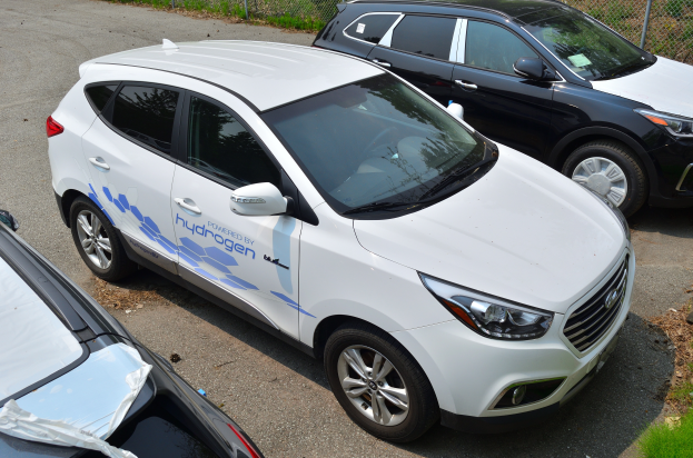 Drei Autos auf einem Parkplatz nebeneinander geparkt, mit einem Maschendrahtzaun und Gras im Hintergrund, eines davon mit der Aufschrift Wasserstofffahrzeug.