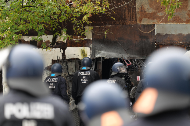 Gruppe von Polizisten in schwarzen Uniformen und Helmen vor einem mit Maschendrahtzaun umgebenen Geb├Ąude mit B├Ąumen im Hintergrund.