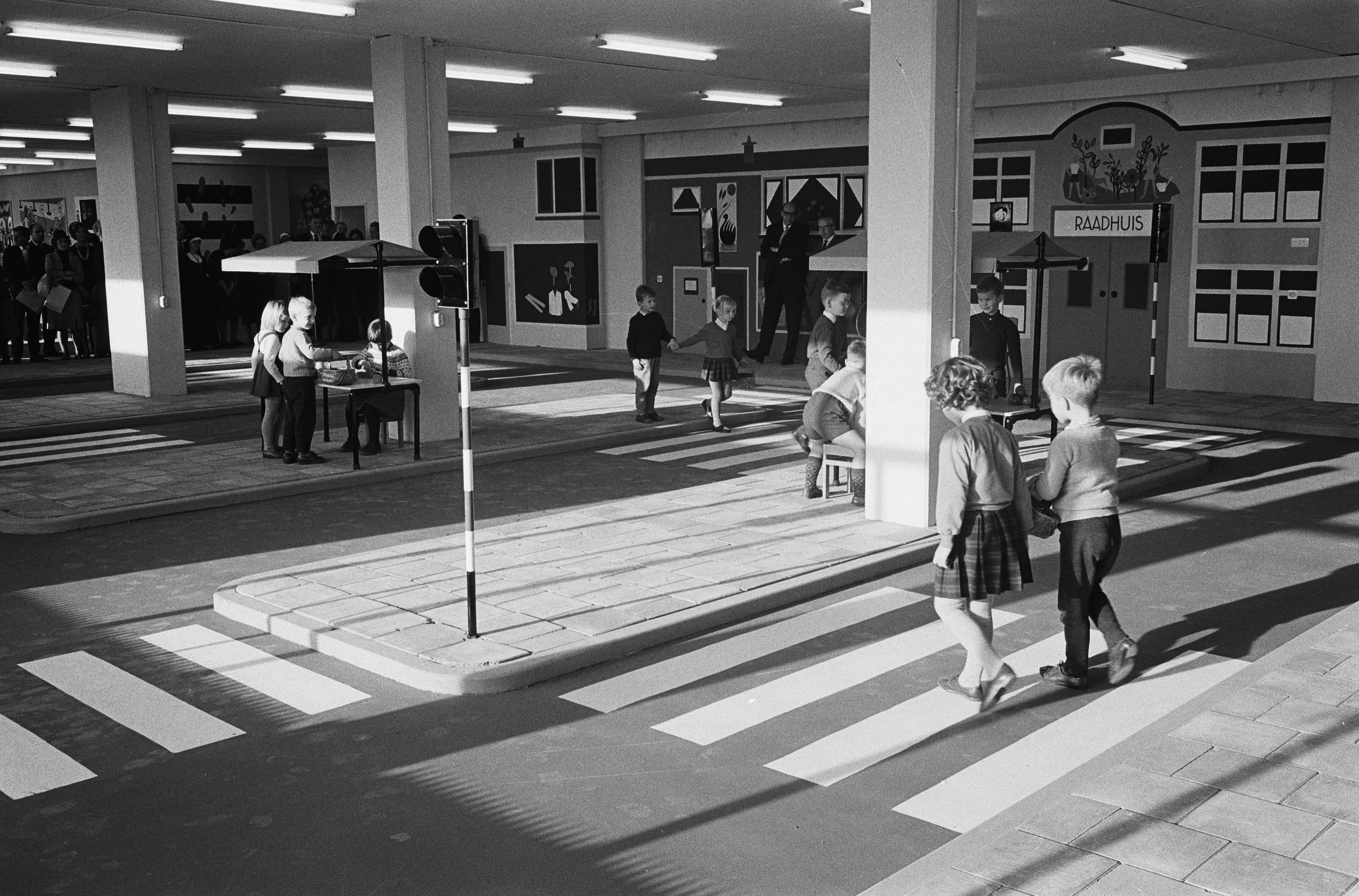 Schwarz-weißes Foto von Kindern, die eine Straße an einer Ampel überqueren, mit Menschen auf Bänken, einem Verkehrssignalmast im Vordergrund und Säulen mit Deckenleuchten im Hintergrund.