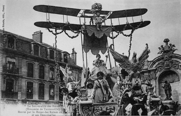 Ein Schwarz-Weiß-Foto eines Umzugswagens mit Statuen und einem Flugzeug, der eine Gruppe von Menschen trägt, mit einem Gebäude und Himmel im Hintergrund und Text unten, der es als Foto des Pariser Karnevals ausweist.