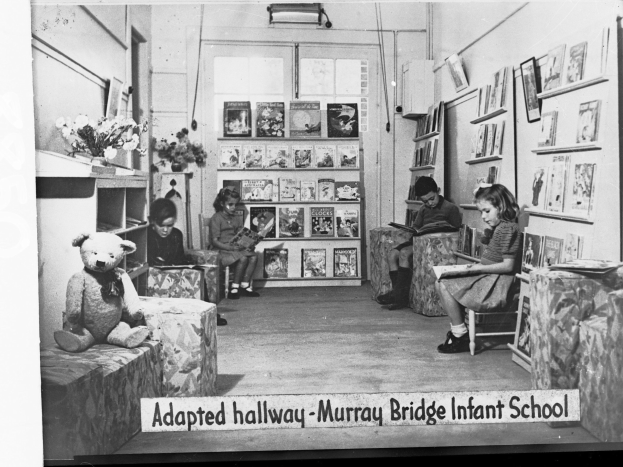 Eine Gruppe von Kindern sitzt auf Stühlen in einem Raum mit Bücherregalen, einem Teddybär, einer Blumenvase und gerahmten Gegenständen an der Wand, mit dem Text "Angepasster Flur Murray Bridge Kindergarten" unten.